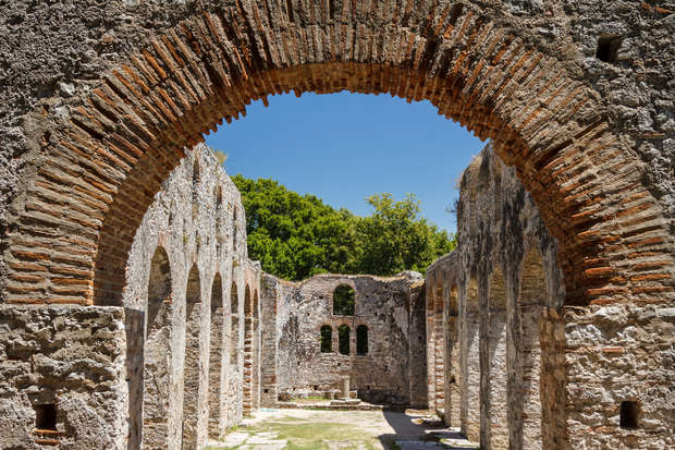 Stone archway frames a view of ancient ruins with tall, textured walls and arched windows. Sunlight illuminates the interior, surrounded by lush greenery and a bright, clear sky.